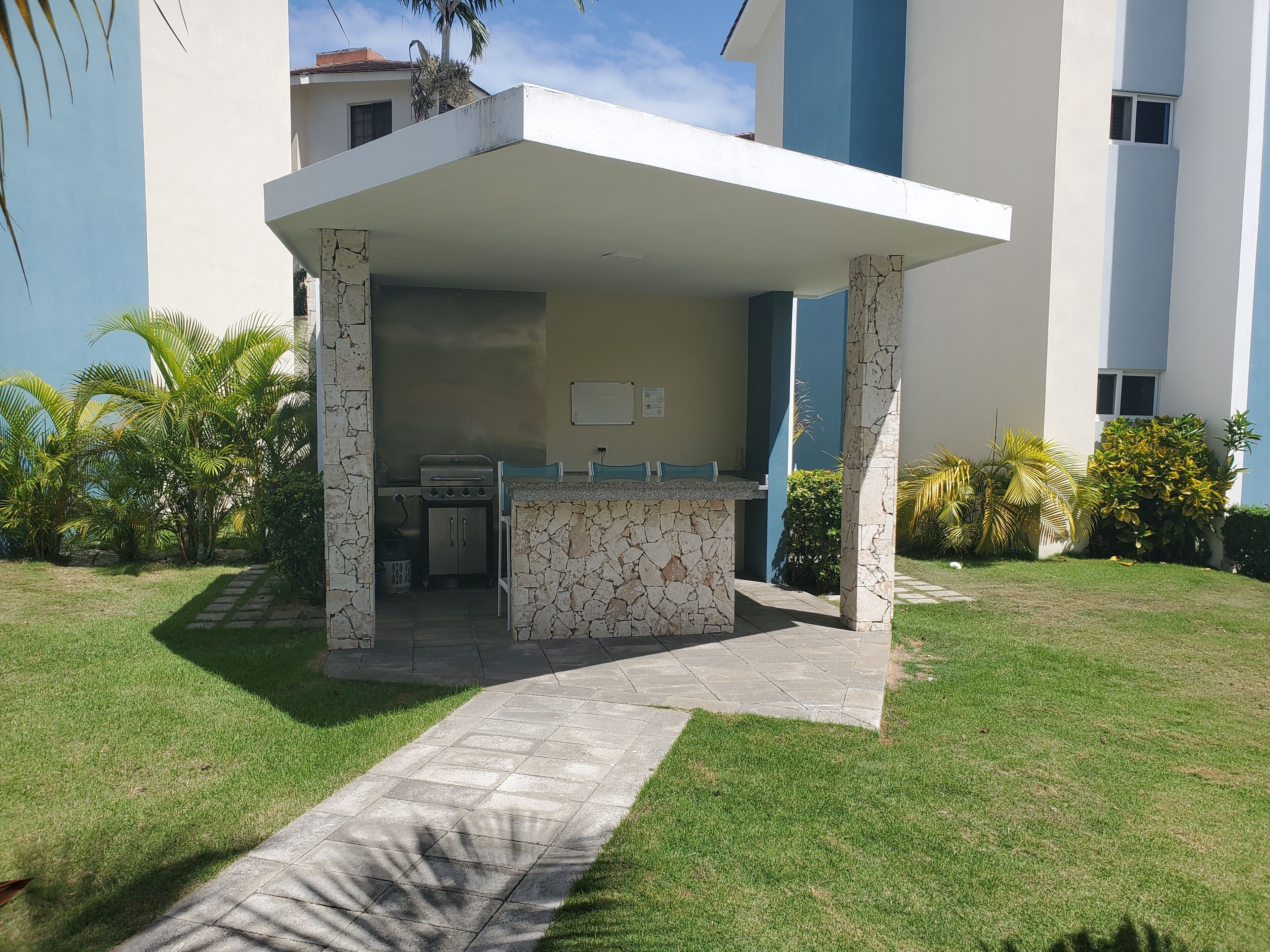 Un'area esterna coperta con un barbecue in pietra e una piccola cucina, circondata da prato curato e palme.