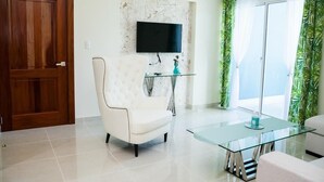 Una sala moderna con un sillón blanco, una mesa de centro de vidrio, un televisor de pantalla plana montado en una pared texturizada y un ventanal corredizo con cortinas verdes.