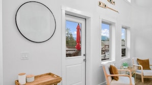 A room with a white door, a round mirror, a wooden side table with jars, two wicker chairs with cushions, and a small table with a plant. - Hideaways| Mins to Lake| King Bed |Dbl Garage| AC (Invermere)