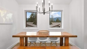 A dining area with a wooden table, three chairs, a chandelier, and a window with a view of trees and mountains. - Hideaways| Mins to Lake| King Bed |Dbl Garage| AC (Invermere)