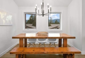 Un comedor con una mesa de madera, tres sillas, una lámpara de araña y un ventanal con vistas a árboles y montañas.