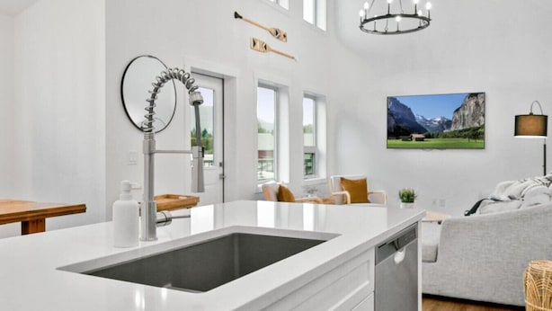 A modern kitchen with a large sink, a dining area with a wooden table, and a living room with a sofa and a painting of mountains. - Hideaways| Mins to Lake| King Bed |Dbl Garage| AC (Invermere)