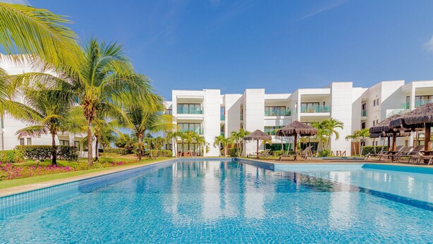 Una piscina rodeada de edificios blancos, palmeras y sombrillas de paja.