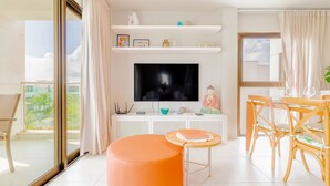 Una sala de estar moderna con un televisor de pantalla plana grande, un comedor con sillas de madera y un balcón con vistas a palmeras.