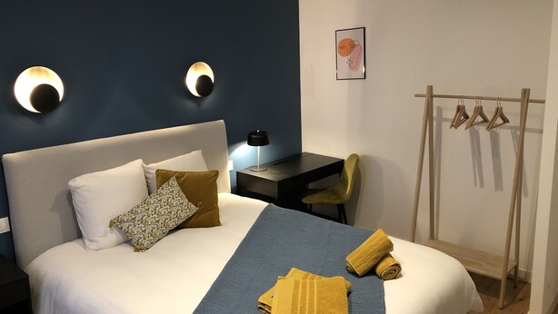 Ein ordentlich bezogenes Bett mit blauer Bettdecke und zwei gelben Handtüchern, ein Schreibtisch mit Lampe, ein Stuhl und ein Kleiderständer mit Kleiderbügeln.