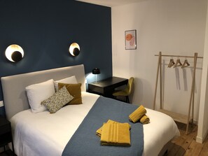 Ein ordentlich bezogenes Bett mit blauer Bettdecke und zwei gelben Handtüchern, ein Schreibtisch mit Lampe, ein Stuhl und ein Kleiderständer mit Kleiderbügeln.
