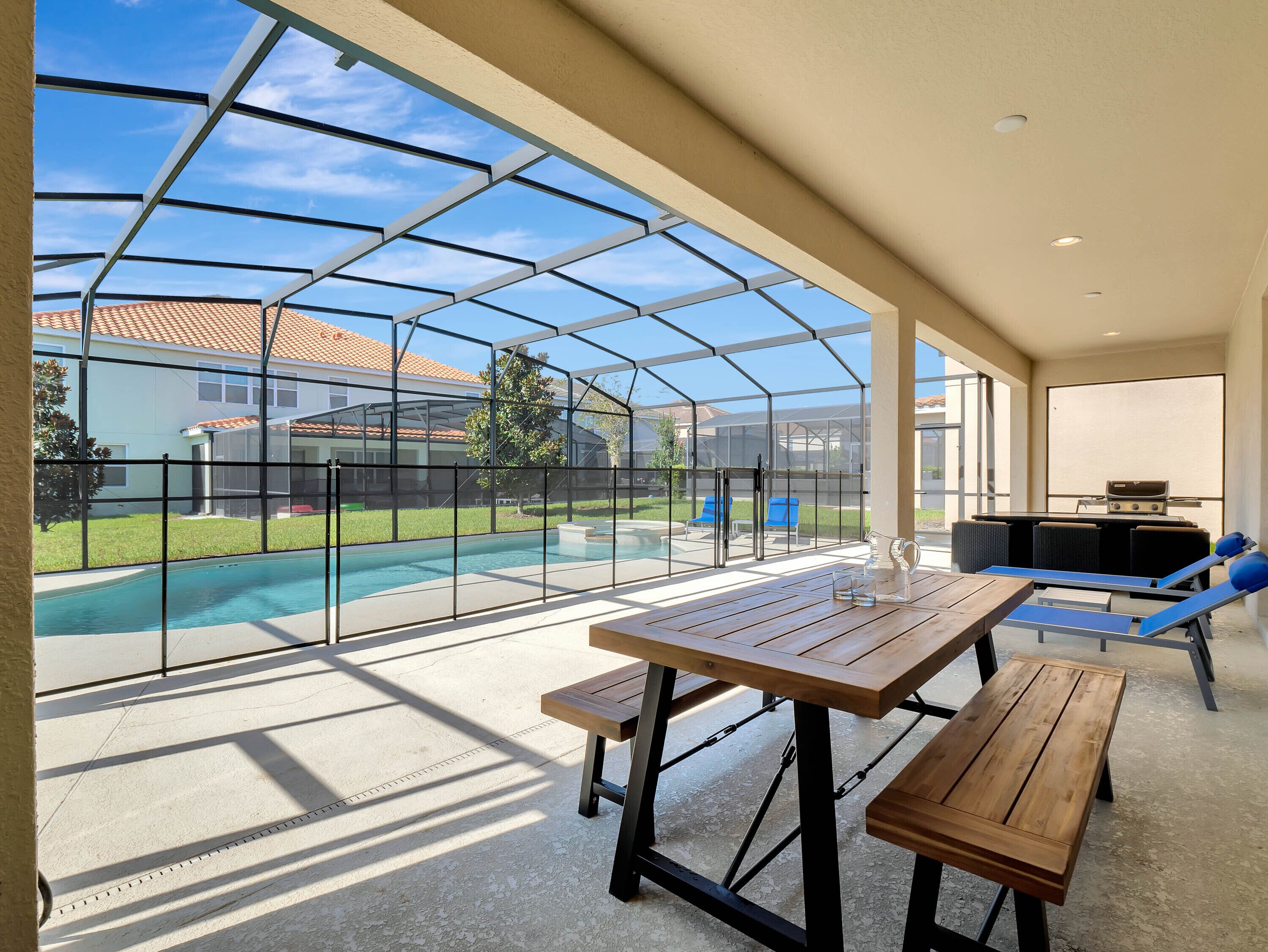 A covered outdoor area with a pool, wooden tables, and chairs, and a barbecue grill.