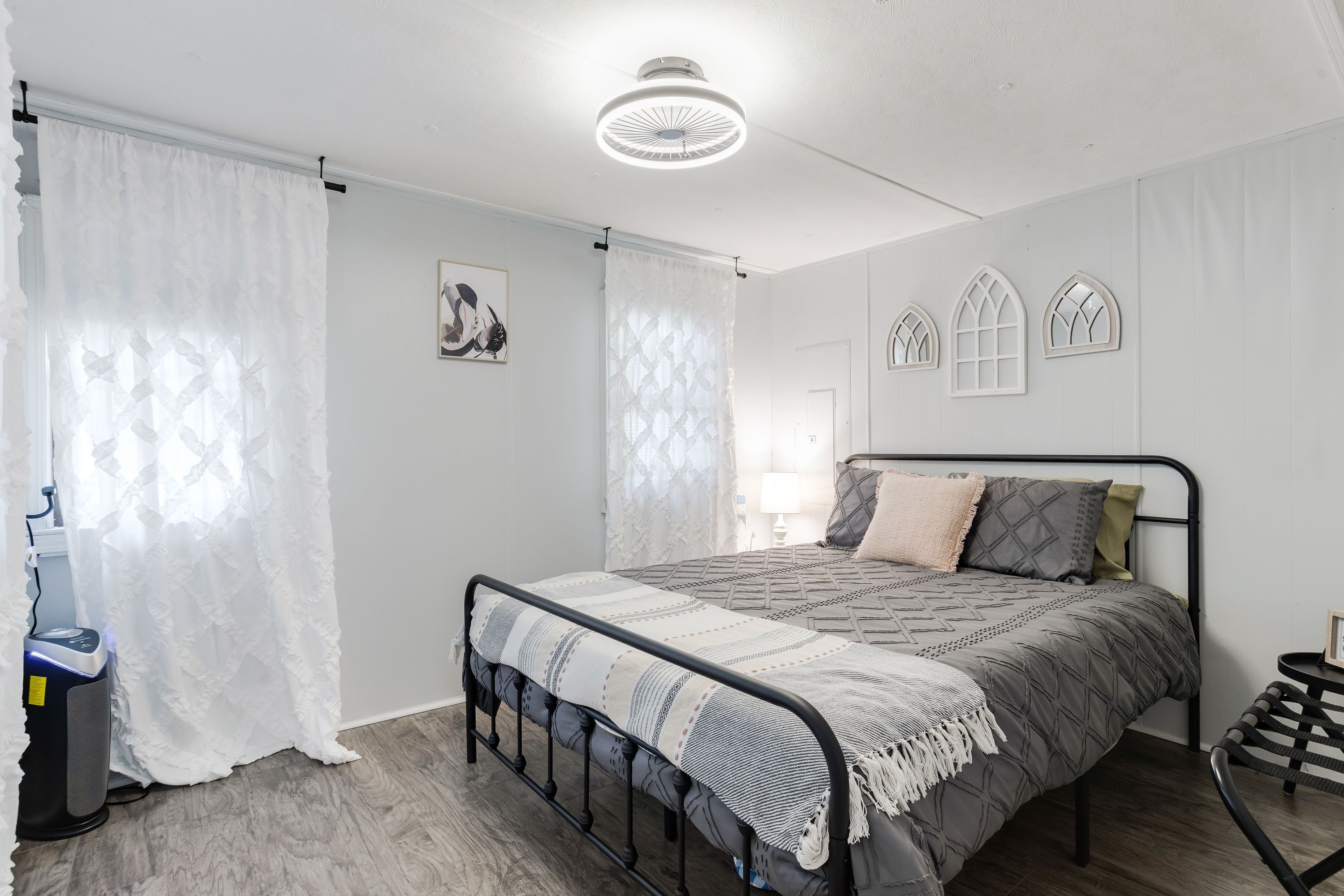 A bedroom with a bed, a bedside table, a chair, a vacuum cleaner, and a window with curtains.