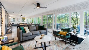 Ein großzügiger Wohnbereich mit grauem Sofa, grünen Sesseln und einem weißen Couchtisch. Große Fenster bieten einen Ausblick ins Freie.