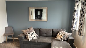Ein Wohnzimmer mit einem grauen Ecksofa, gemusterten Kissen, einem blauen Teppich und einem Spiegel an der Wand.