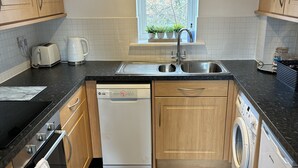 Eine kleine Küche mit Holzausstattung, einer Spülmaschine, einer Waschmaschine und einem Fenster mit Jalousien.