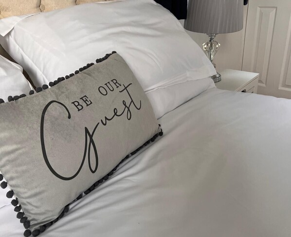 Una cama con ropa blanca y una almohada con la frase "BE OUR Guest".