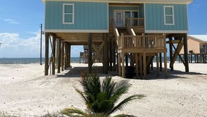 A two-story wooden beach house with a balcony, situated on a sandy beach with a clear view of the ocean. - Toes in the Sand - Gulf Beachside, 4 Bedroom, 4 Bath, Dog Friendly, Sleeps 12 (Dauphin Island)