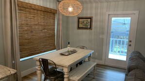 Un comedor con una mesa de madera, sillas, un banco, una lámpara colgante, una ventana con persianas, un cuadro enmarcado y una puerta que da al exterior.