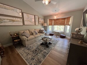 A living room with a beige sofa, a glass coffee table, a patterned rug, wooden flooring, and a large window with blinds.