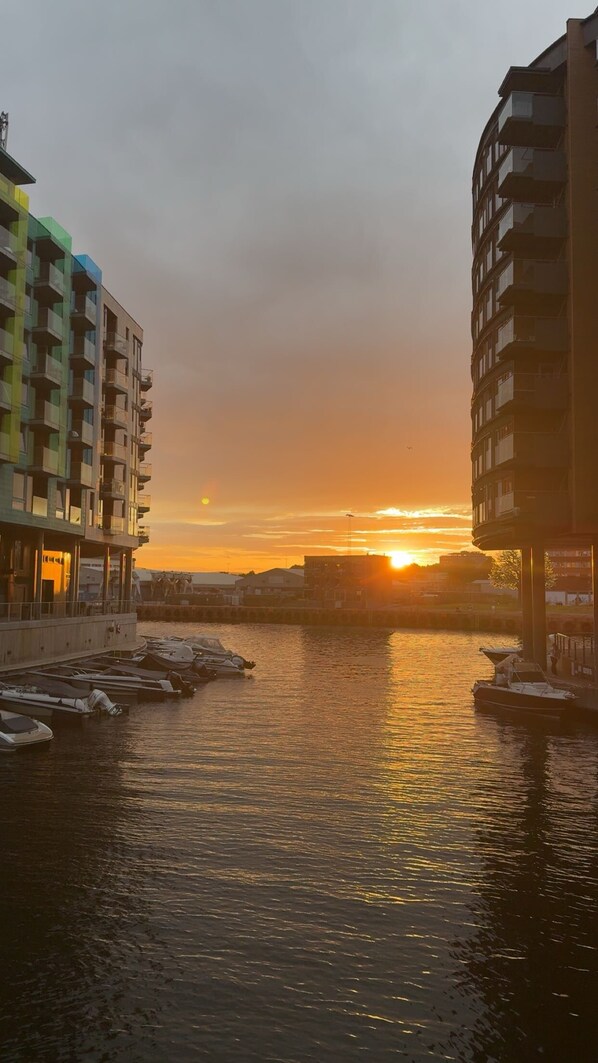 Vista al agua con edificios a ambos lados, un atardecer en el fondo y barcos amarrados a lo largo de la orilla.