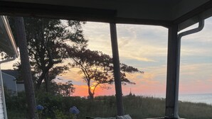 A room with a sofa and a glass coffee table, a view of trees and a sunset.