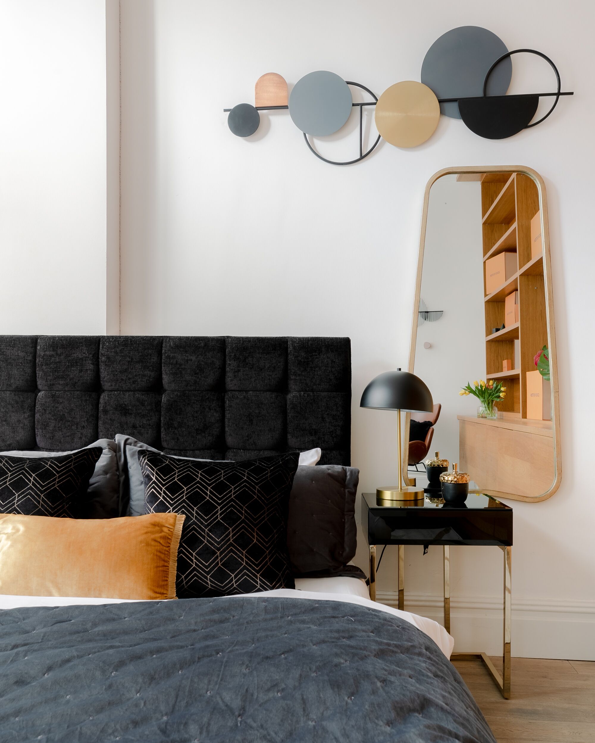 A modern bedroom with a tufted headboard, a black and gold bedside table, a mirror, and a wall art piece.