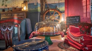 Un comedor con una mesa dispuesta para una comida, que incluye un recipiente tapado, una rueda de barco y un letrero en una pizarra.