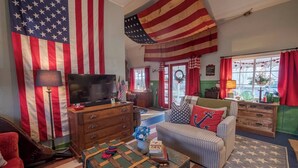 A living room with a large American flag, a television, a couch with nautical-themed pillows, and a vintage suitcase.