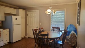 A kitchen with wooden cabinets, a refrigerator, a dining table with chairs, and a window with blinds. - Peggy  Sue (Pass Christian)