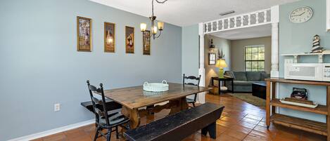 A dining area with a wooden table, chairs, and a bench. There is a chandelier hanging from the ceiling, and a clock on the wall.