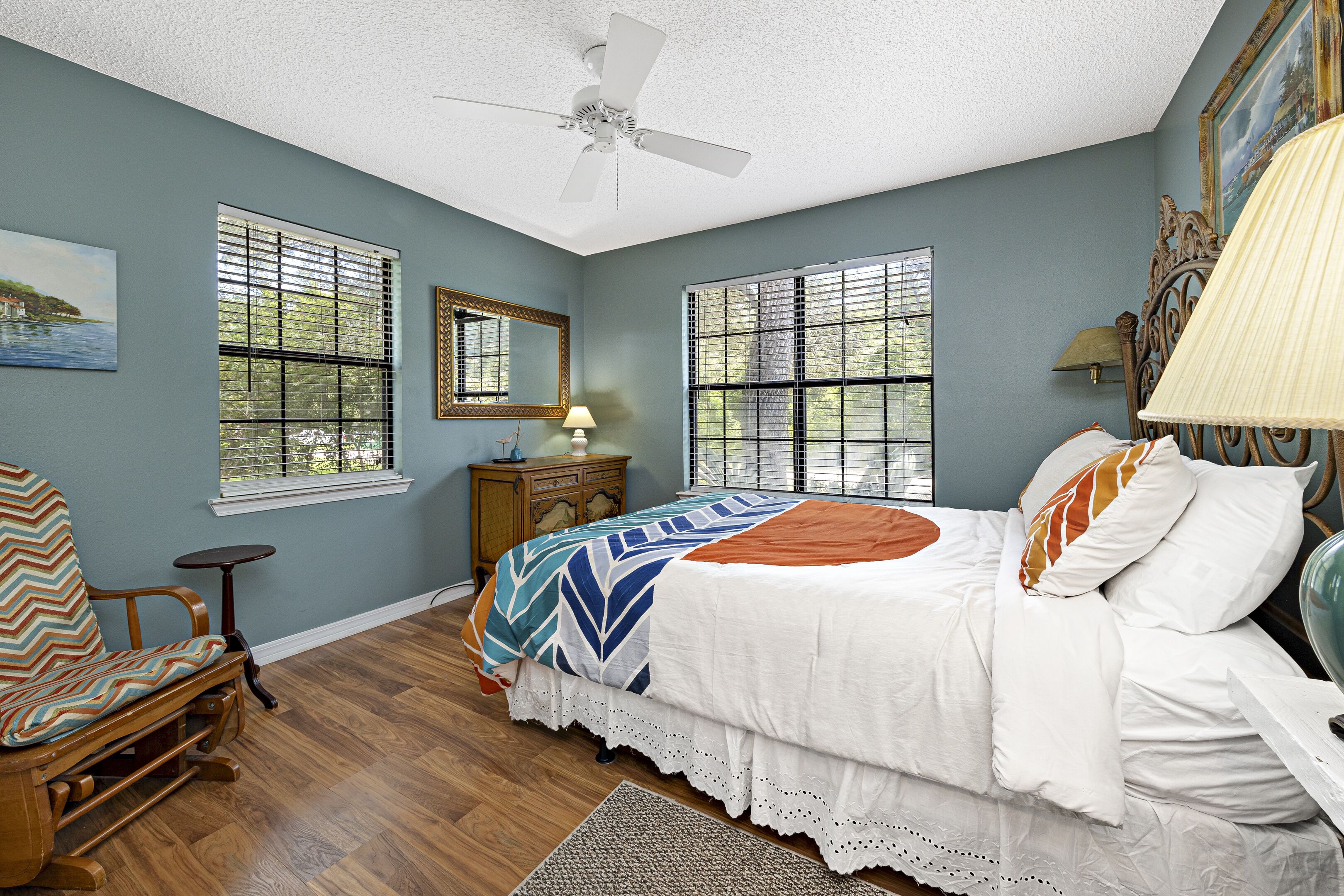 A bedroom with a bed, a chair, a nightstand, a lamp, and two windows with blinds.