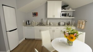 Una cocina moderna con armarios blancos, un refrigerador blanco, un microondas y una cafetera sobre la encimera. Hay una mesa de comedor con sillas y un florero con flores.