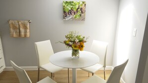 Un comedor con una mesa redonda, cuatro sillas blancas, un jarrón de flores, una lámpara en el techo y un cuadro en la pared.