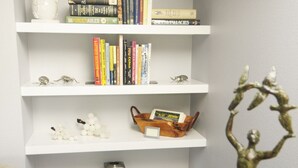 Una habitación con una estantería blanca llena de libros, un pequeño jarrón blanco y una escultura decorativa sobre una mesa de madera.