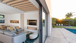 Uma sala de estar moderna com sofá, mesa de centro e televisão. Há uma porta de vidro que dá para uma área de piscina com mobiliário de exterior.