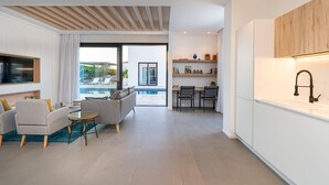 Uma sala de estar moderna com sofá, mesa de centro e televisão. Há uma cozinha com pia e armários. Uma piscina é visível pelas portas de vidro.