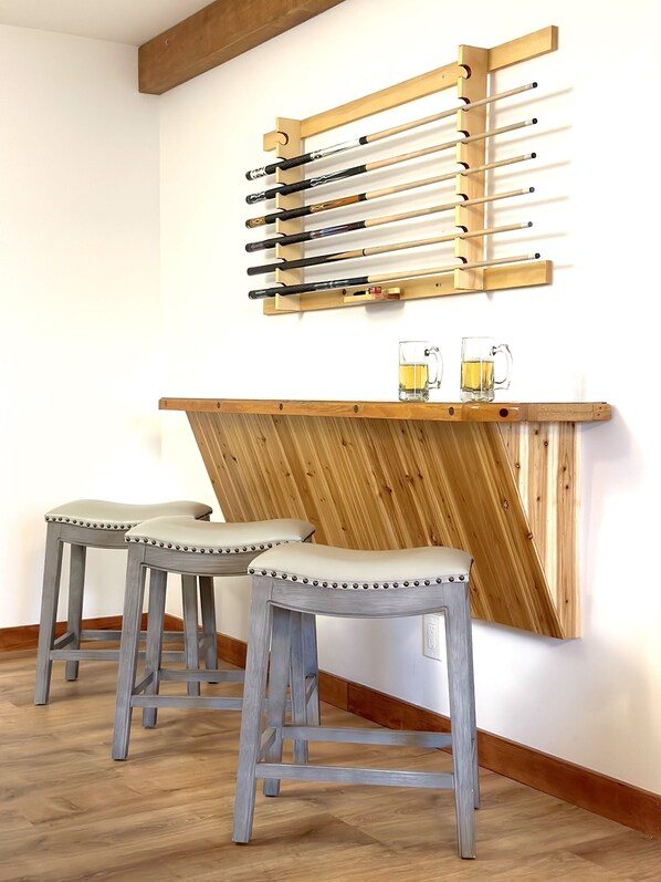 A bar area with two stools, a wooden bar counter, and a wall-mounted pool table rack. - Superb Lodge w Heated Pool, Large Hot Tub, Game Room, 15mins to Gore Mountain! (Johnsburg)