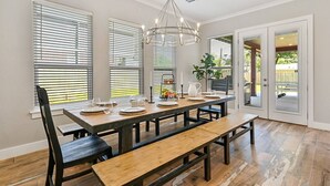 A dining area with a long wooden table, black chairs, and benches. There is a chandelier above the table, and the room has large windows with blinds. - Aggieland Crossing ~ Huge Gameday Backyard! (College Station)
