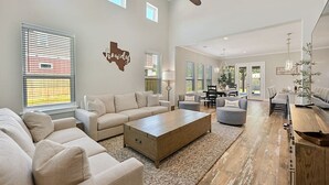 A spacious living room with a large ceiling fan, beige sofas, a wooden coffee table, and a dining area with a chandelier. - Aggieland Crossing ~ Huge Gameday Backyard! (College Station)
