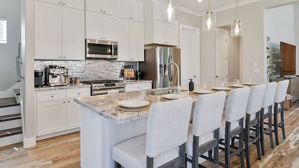 A modern kitchen with white cabinets, stainless steel appliances, a large island with bar stools, and pendant lighting. - Aggieland Crossing ~ Huge Gameday Backyard! (College Station)