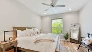 A bedroom with a bed, bedside table, nightstand, mirror, and a window with blinds. - Aggieland Crossing ~ Huge Gameday Backyard! (College Station)