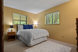 A bedroom with a bed, nightstand, lamp, and two windows with blinds.