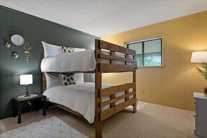 A bunk bed in a room with a dark green wall, a small bedside table with a lamp, and a window with blinds.