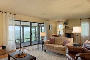 A living room with a sofa, a coffee table, and a fireplace. There is a large window with curtains and a view of the outdoors.