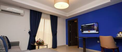 Una habitación de hotel moderna con un sofá gris, una pequeña mesa redonda con un arreglo floral, un televisor en una pared azul y una unidad de aire acondicionado en la pared.