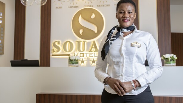 A hotel reception area with a modern design, featuring a reception desk, a clock, and a sign with the hotel's name and logo. - Souare Premium Hotel (Conakry)