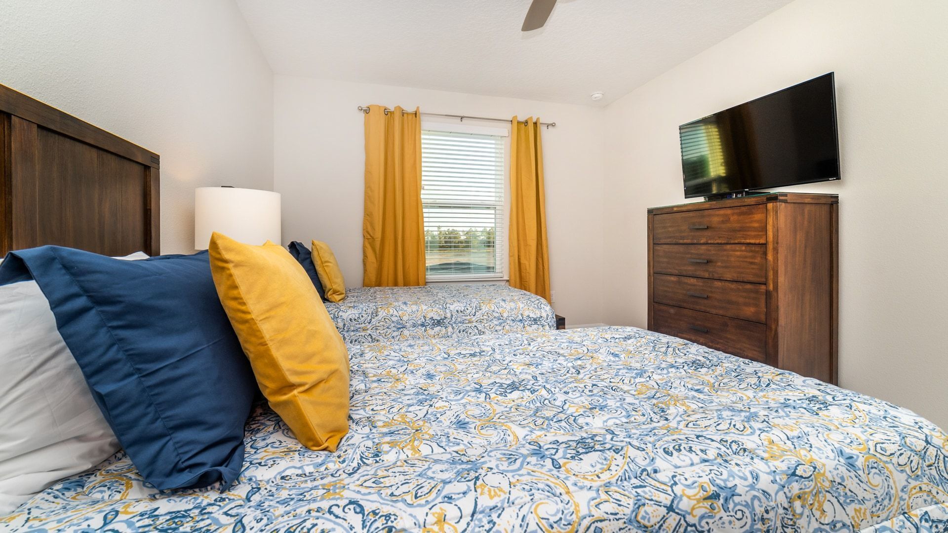 A bedroom with a bed, a wooden dresser, a flat-screen TV, and a window with yellow curtains.