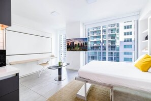 A modern hotel room with a bed, a desk, a chair, a small table with a plant, a TV, and a cityscape view.