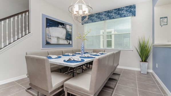 A dining area with a long table set for six, a chandelier, a painting, and a potted plant.