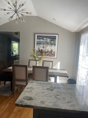 Un coin repas doté d’un plan de travail en marbre, de chaises en bois, d’un tableau représentant une scène de plage et d’un lustre moderne.
