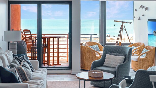 A modern living room with a large sliding glass door leading to a balcony with a view of the beach. There are wicker chairs, a sofa, and a small table. - Coastal Haven by Gulf and Sound (Pensacola Beach)
