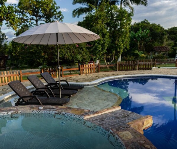 Una piscina con sillas de descanso y una sombrilla, rodeada por una valla de madera y abundante vegetación.
