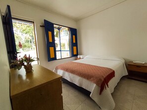 Un dormitorio con cama, mesita de noche, cómoda de madera y ventana con contraventanas azules.
