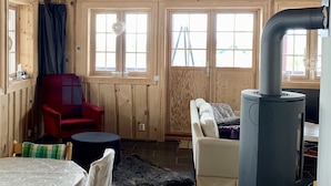 Interior acogedor de una cabaña con paredes de madera, una mesa de comedor con sillas, un sofá, una silla roja, un calentador negro y una ventana con cortinas.